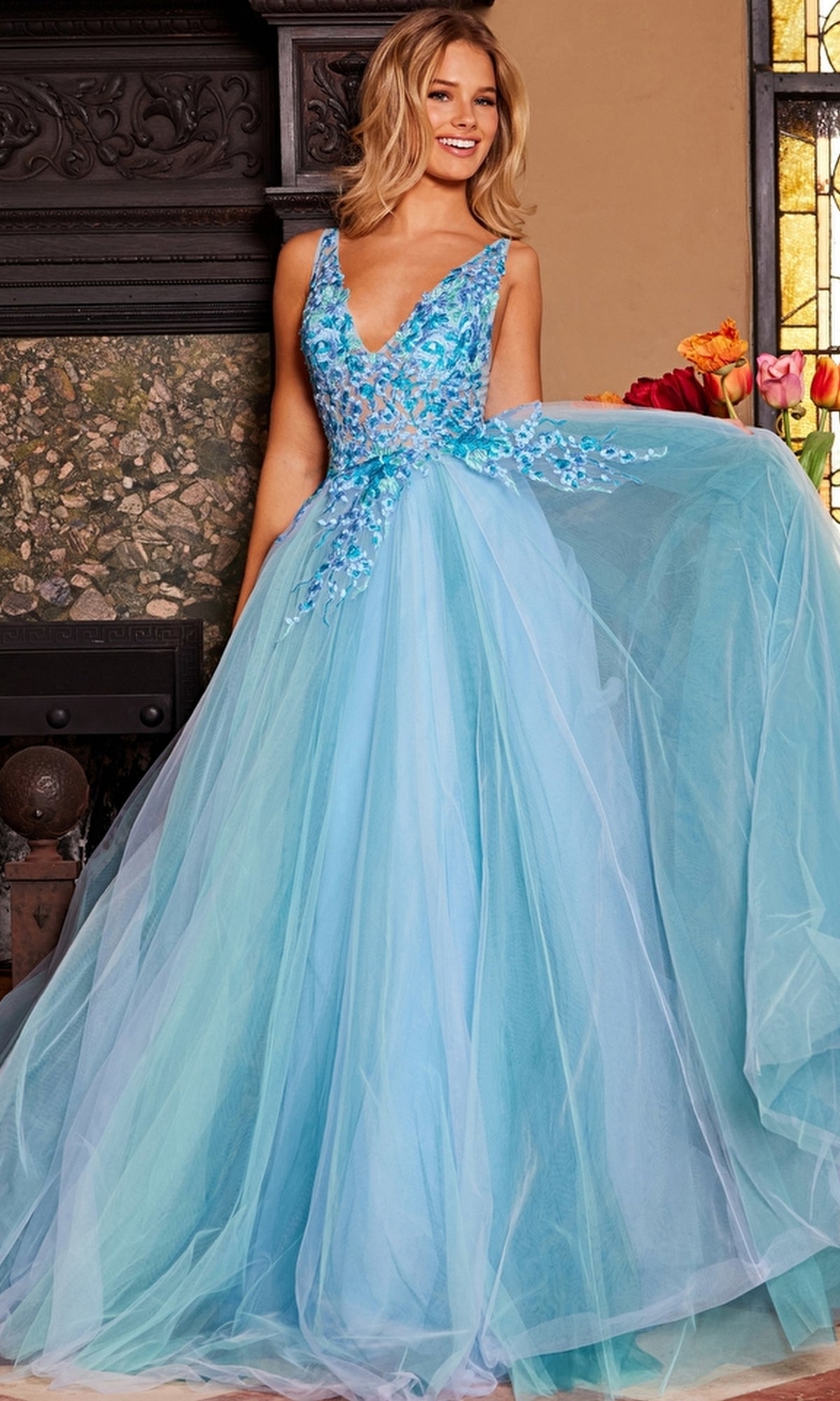 A woman stands indoors, gracefully holding out one side of her Jovani Tulle Prom Ball Gown with Floral Embroidery, featuring a sheer bodice and a flowing light blue tulle skirt.